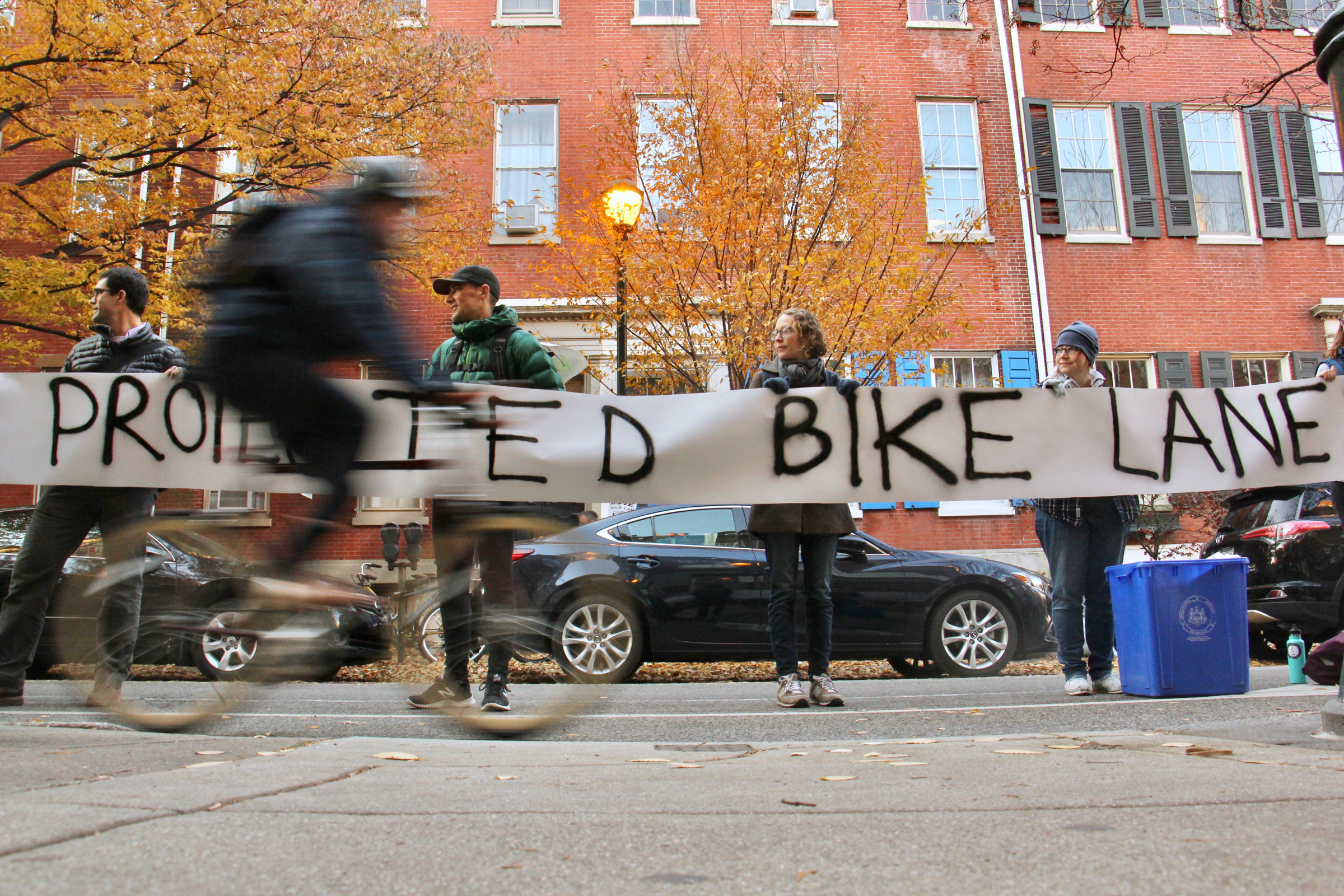 Bicyclists forming a human-protected bike lane hold up a banner calling for protected bike lanes near site of a fatal crash the day prior