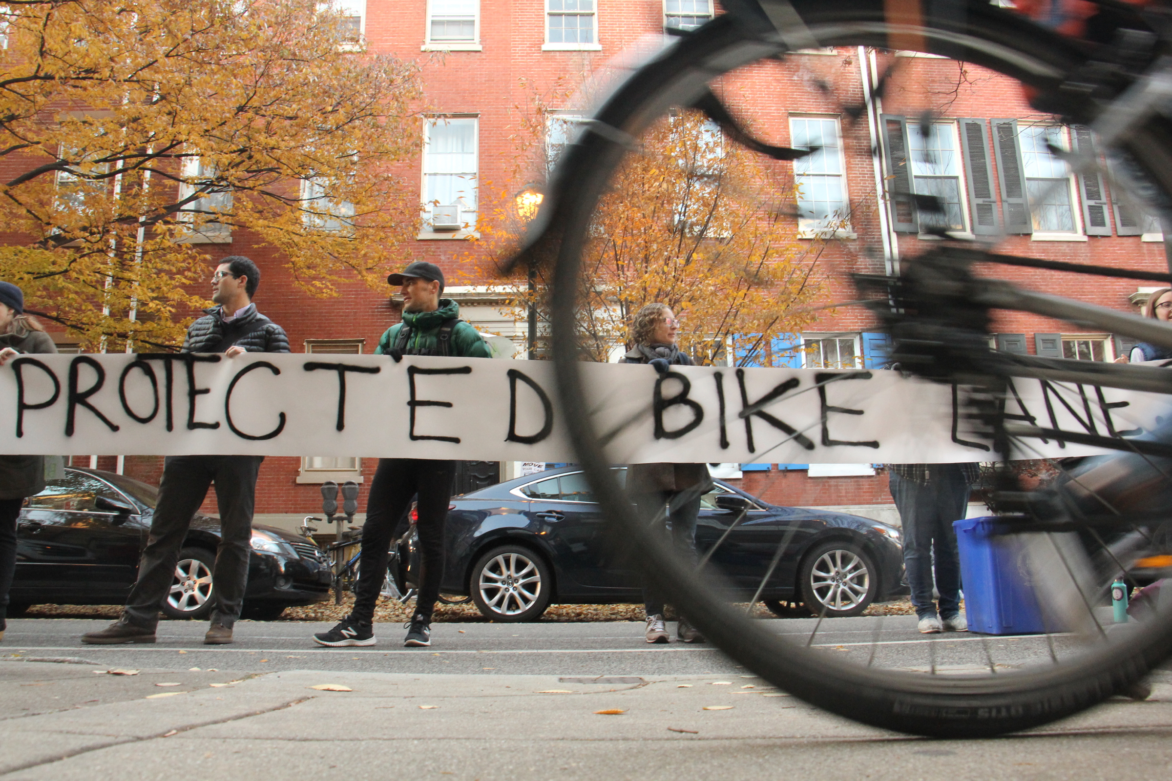 Bicyclists forming a human-protected bike lane hold up a banner calling for protected bike lanes near site of a fatal crash the day prior (Emma Lee, WHYY)