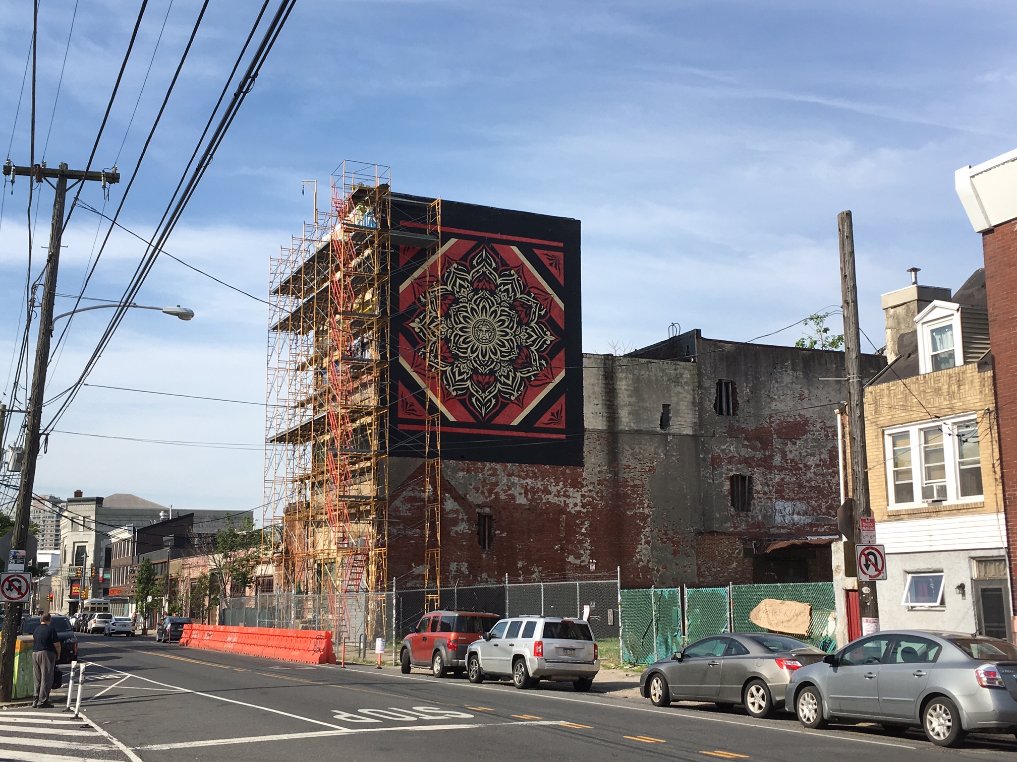 Scaffolding surrounds the proposed site of a Fishtown hotel, next to Frankford Hall. The sidewalk has been blocked off for years. A Shepard Fairey mural is painted on the side of the building. 